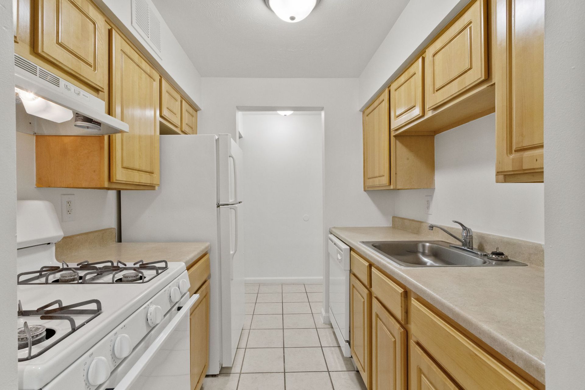 Kitchen with light-colored wooden cabinets, white appliances, and tile flooring. An entryway is visible in the distance.