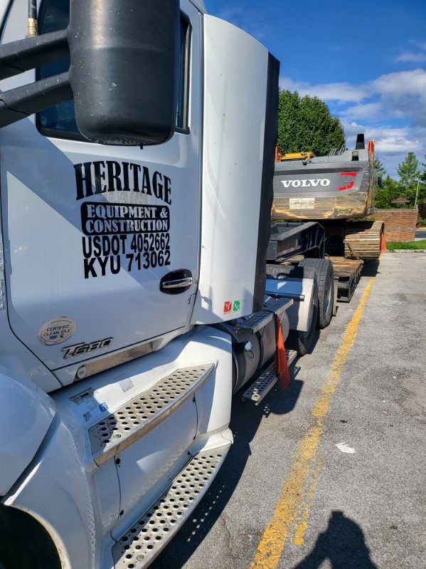 A white semi truck is parked in a parking lot next to a volvo excavator.