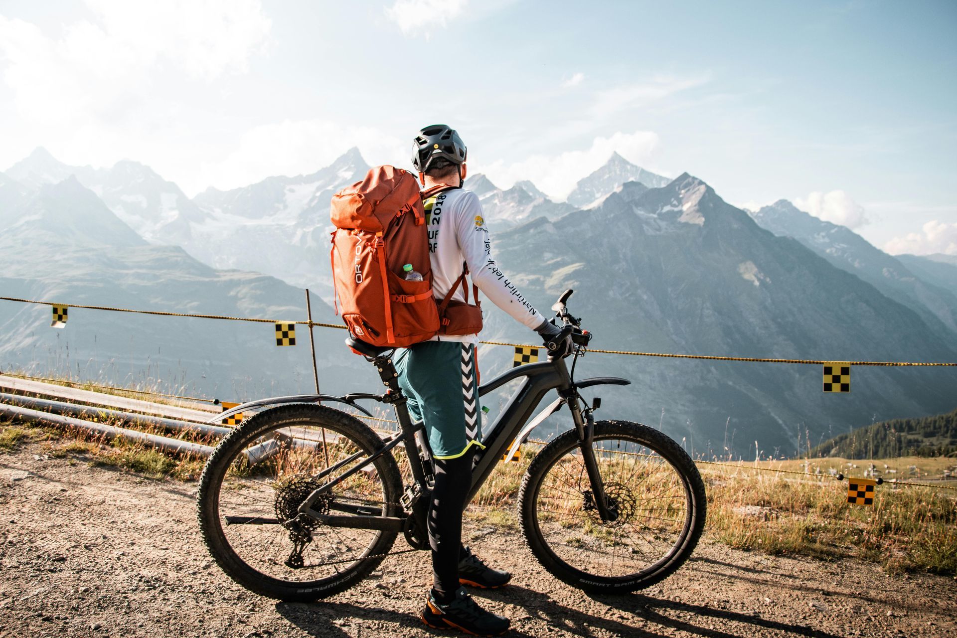 A man with a backpack is standing next to a bicycle on a dirt road.