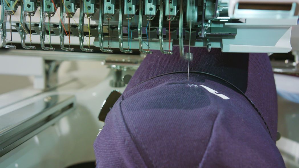 Close-up of a multi-needle embroidery machine stitching a white design onto a dark purple baseball cap.
