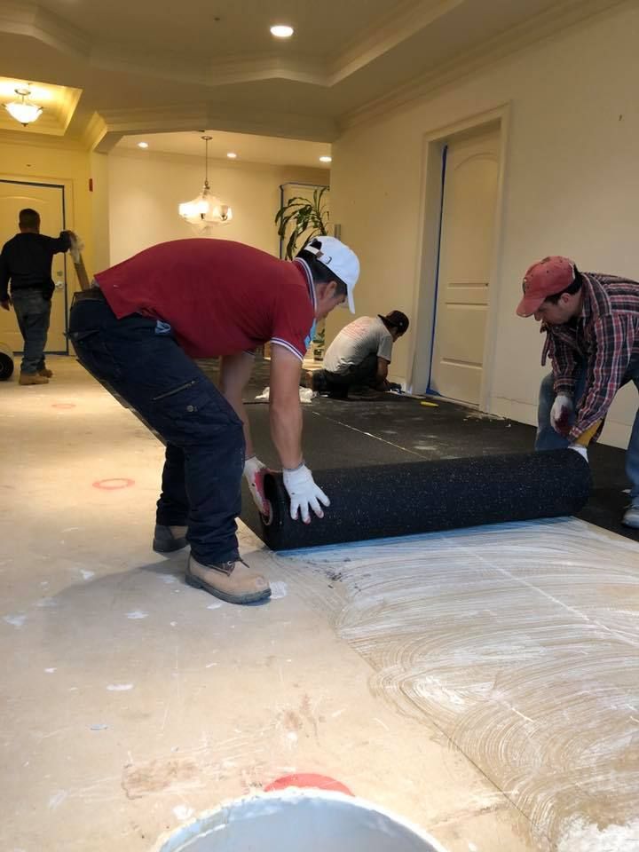 Workers installing flooring, rolling out a black mat on a light-colored surface in an indoor setting.