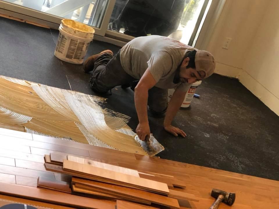 Man kneeling, spreading adhesive for wood flooring installation in a room.