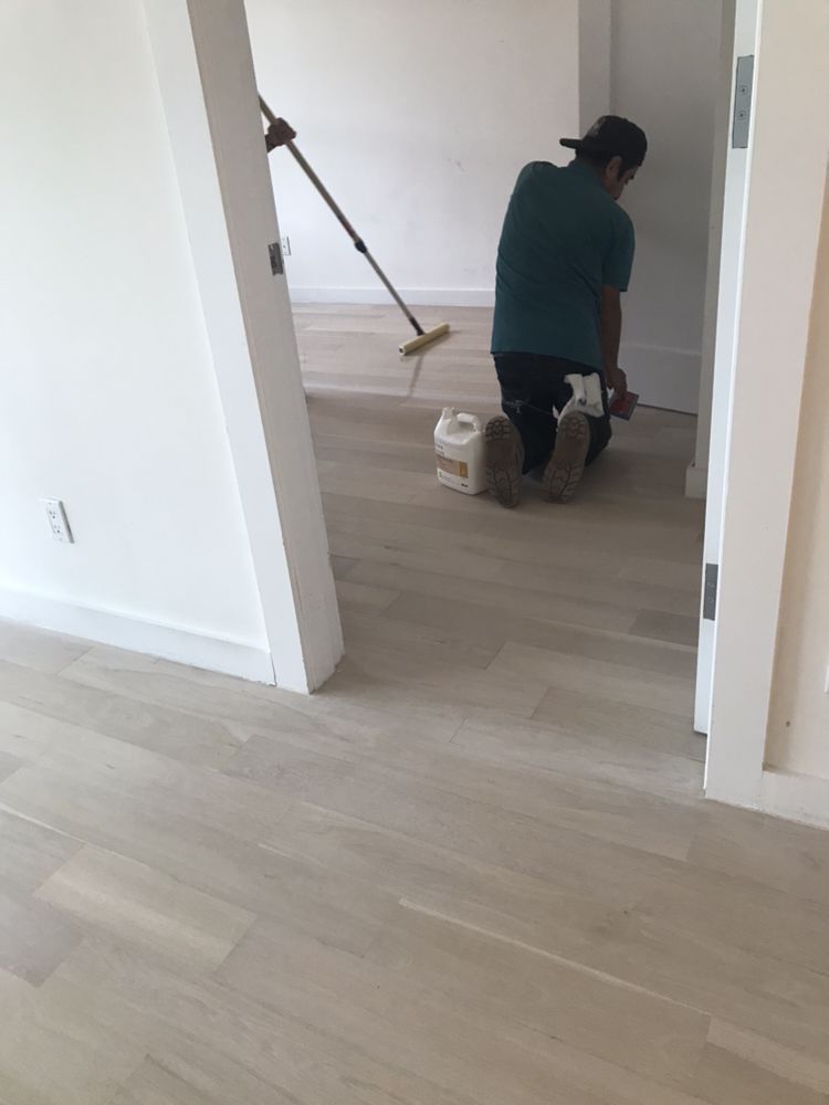 Person applying finish to light-colored hardwood floor in a room, with a second person using a long handled applicator.
