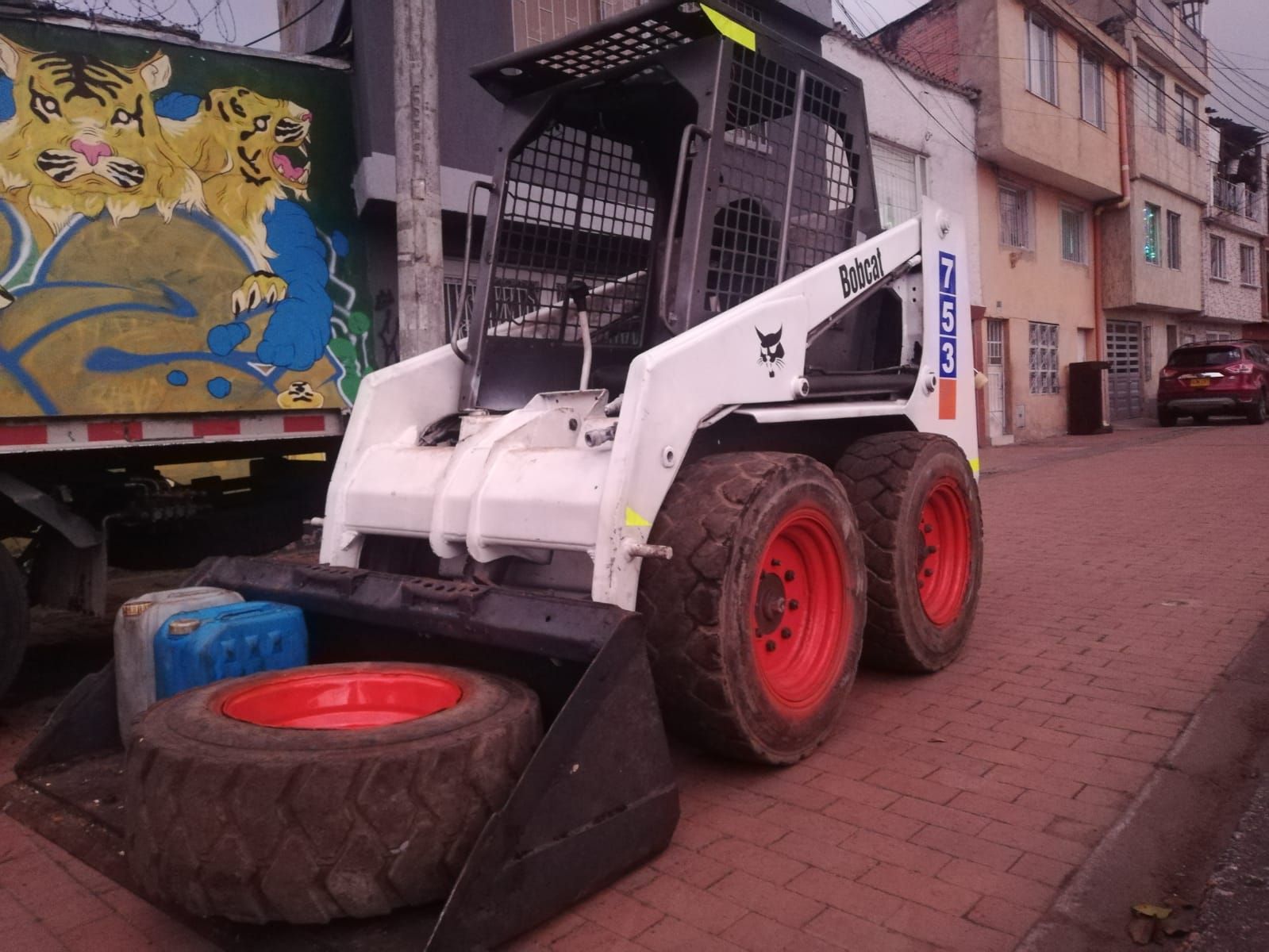 Minicargadora blanca al lado de un camión en una calle estrecha, con un neumático y una rampa negra delante.