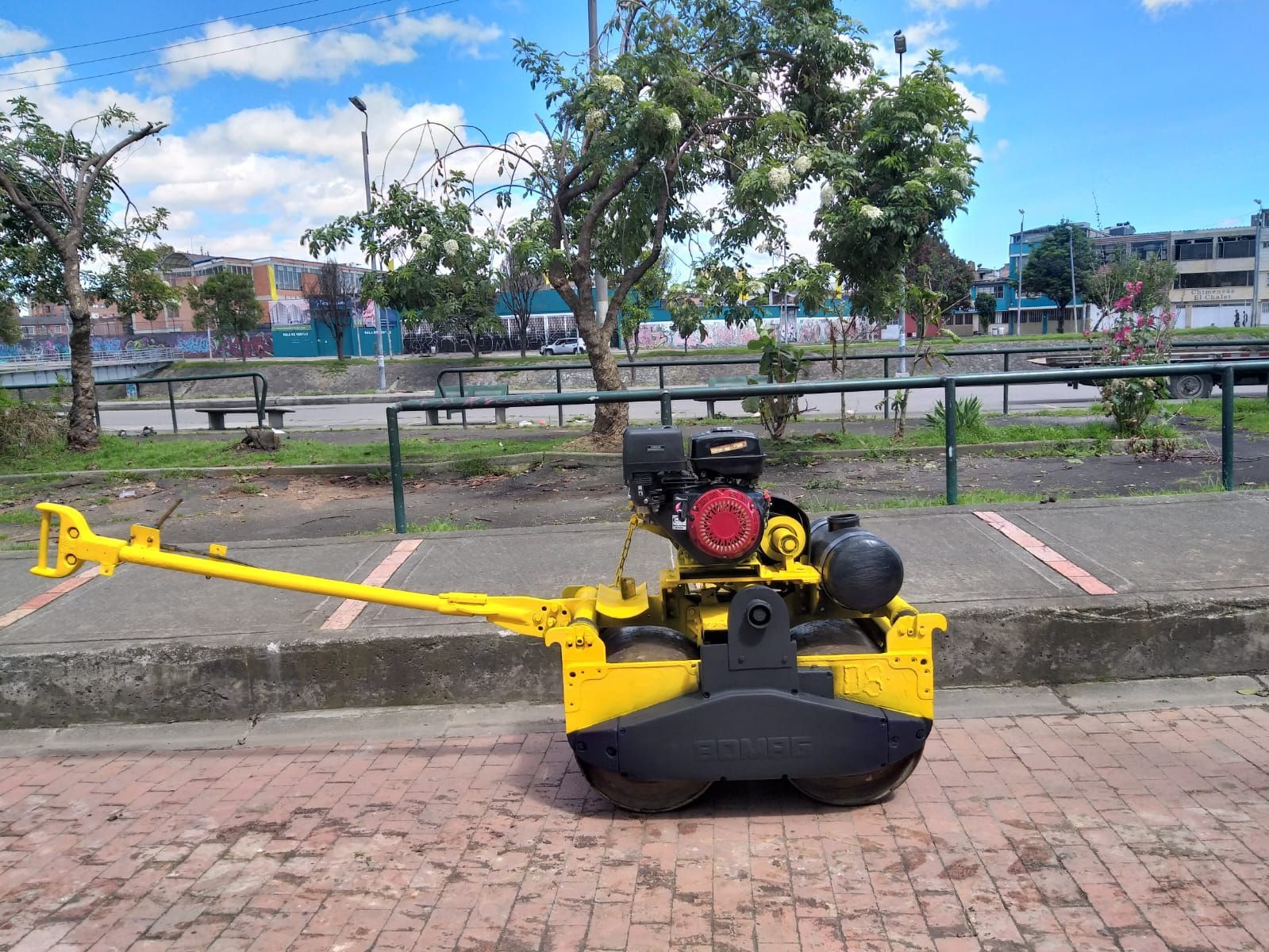 Una apisonadora amarilla estacionada en un camino de ladrillos junto a una calle con árboles y bancos.