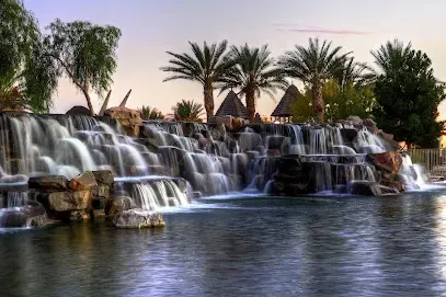 Mobile IV Therapy Las Vegas-A serene waterfall flows over rocks into a calm pool, surrounded by palm trees and pyramidal structures, under a soft pink and blue sky at dusk.