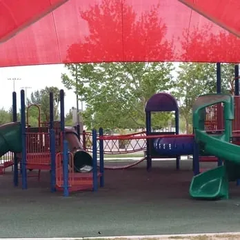 Mobile IV Therapy Las Vegas-Children's playground with red, blue, and green equipment under a red shade. Features slides and climbing structures. Trees in the background. Calm scene.