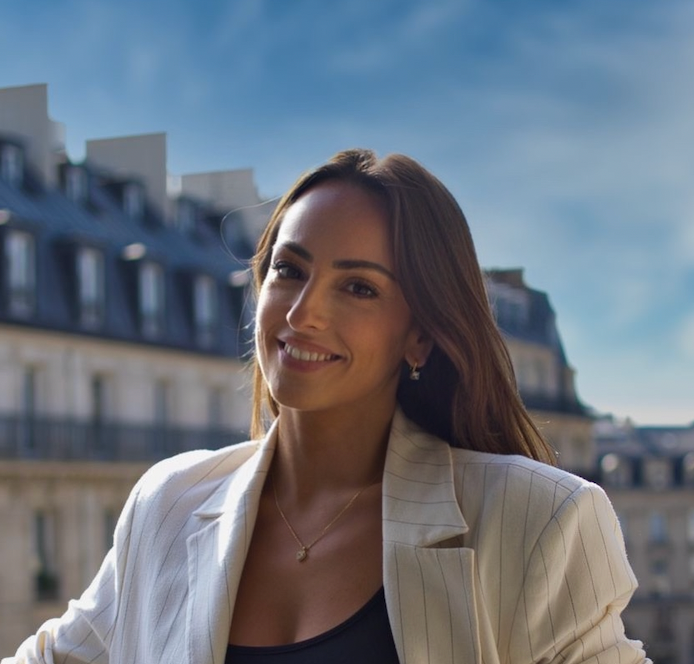 Une femme en blazer blanc sourit à l'extérieur, avec l'architecture parisienne en arrière-plan.