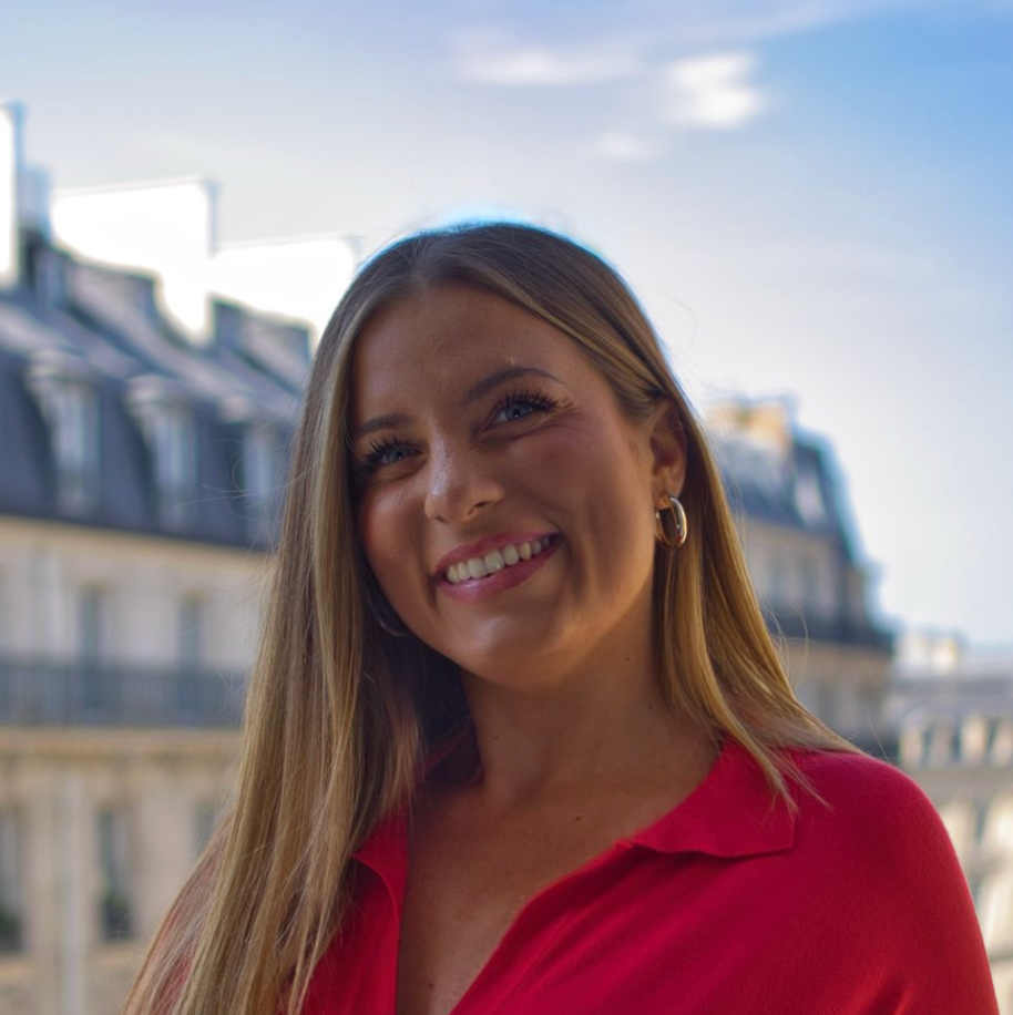 Une femme aux cheveux blonds sourit à la caméra devant un immeuble parisien. Elle porte un haut rouge.