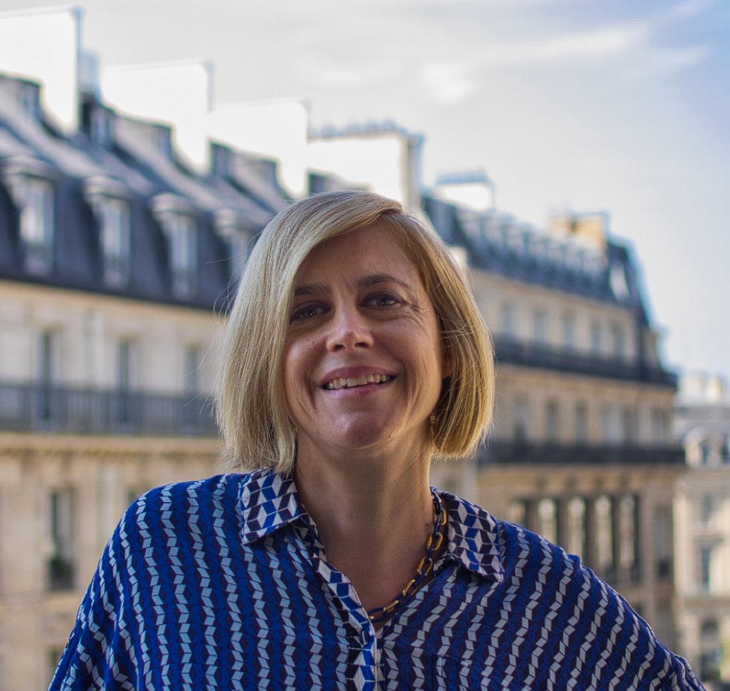Une femme aux cheveux blonds sourit devant des immeubles parisiens, vêtue d'une chemise à motifs bleus.