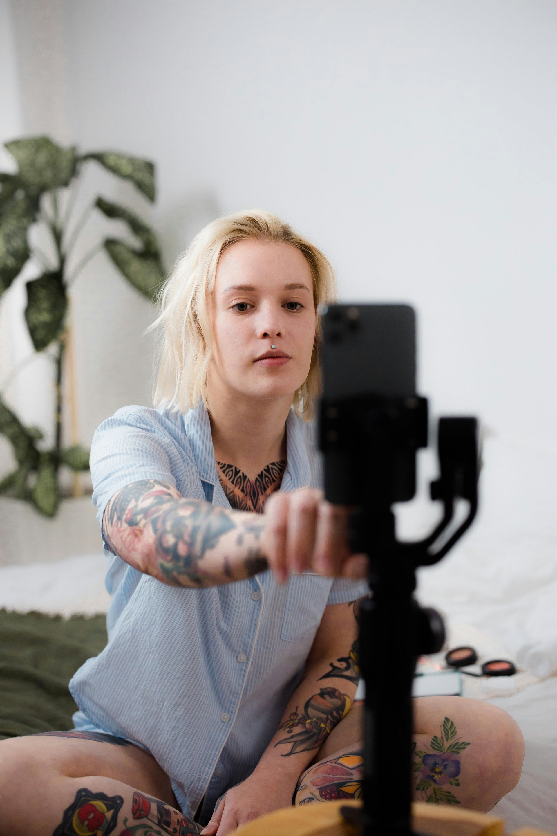 Une personne blonde avec des tatouages ​​​​enregistrant une vidéo avec un téléphone sur un stabilisateur, assise sur un lit.