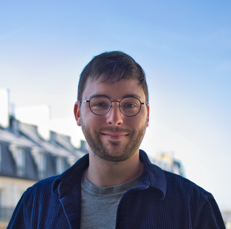 Un homme avec des lunettes sourit, veste bleue, chemise grise, contre les immeubles parisiens.