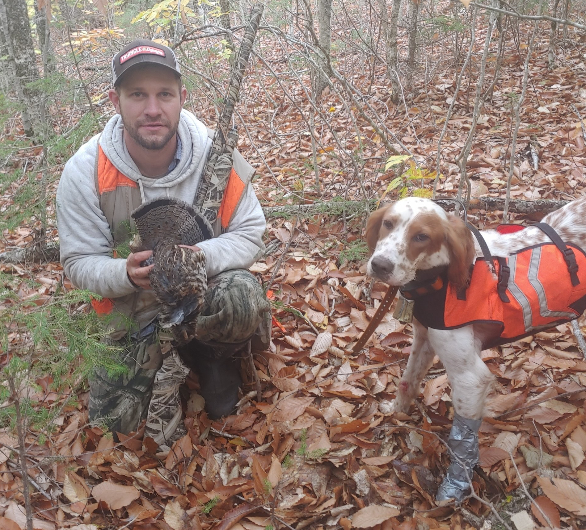A man is holding a bird next to a dog wearing an orange vest.