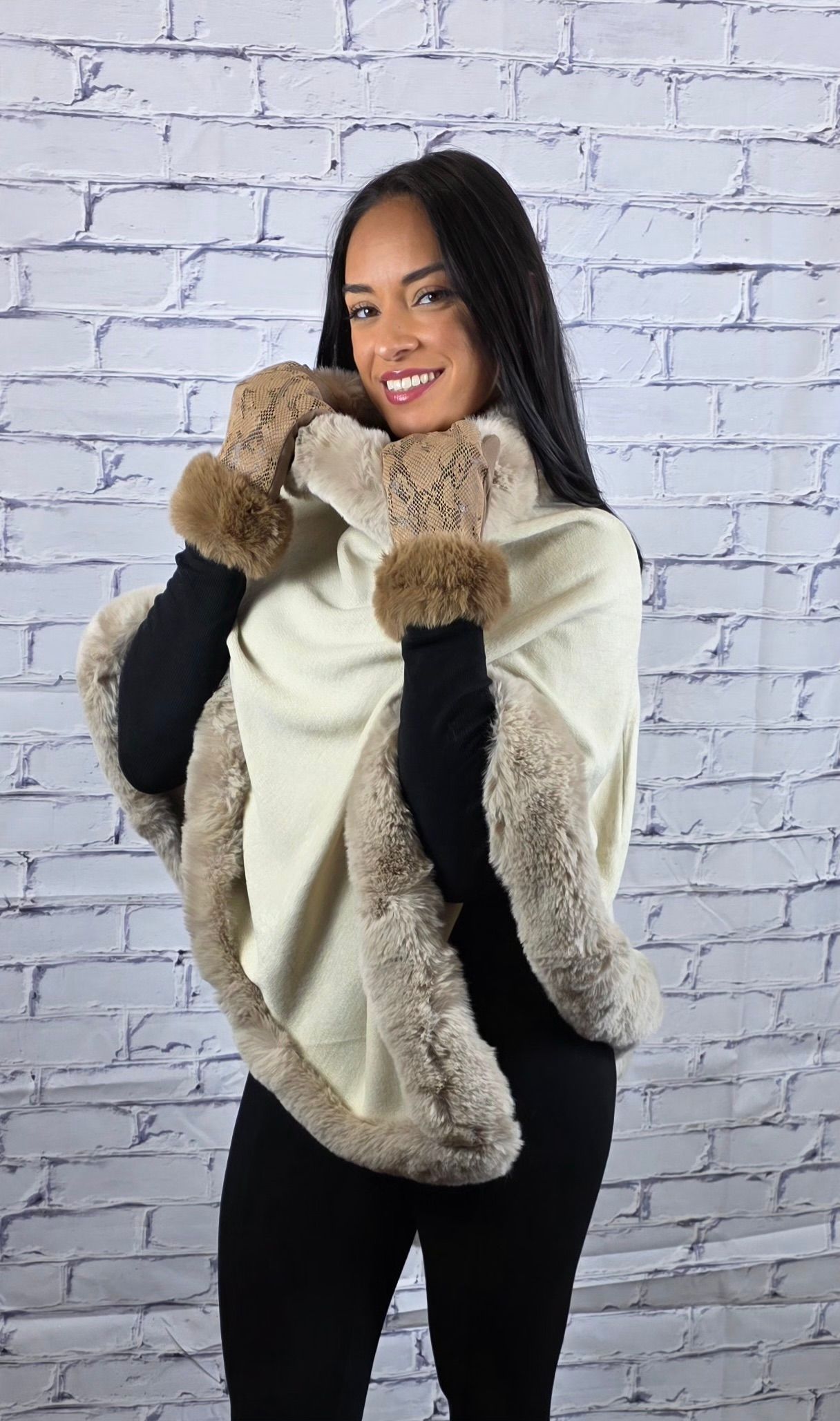 Woman wearing cream-colored shawl with brown fur trim, black sleeves, and black pants, smiling in front of a white brick wall.