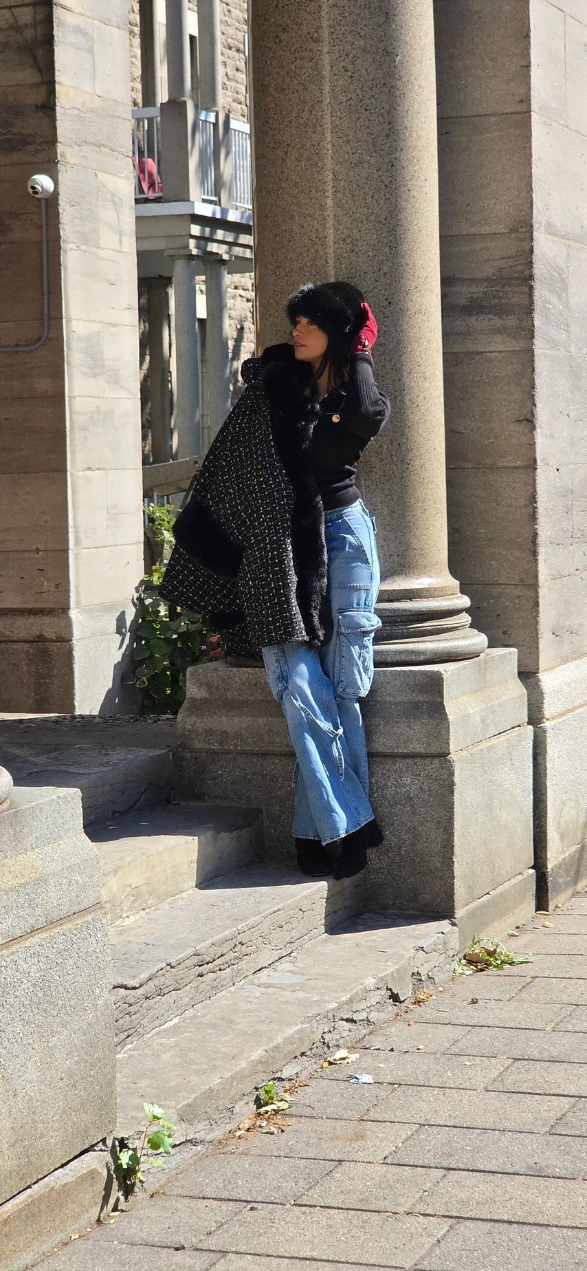 A person leans against a pillar, wearing a black coat, blue jeans, and a red hat, outside.