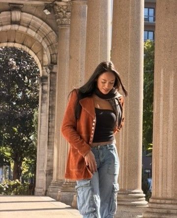 Woman in orange coat and jeans walking under an archway of columns.