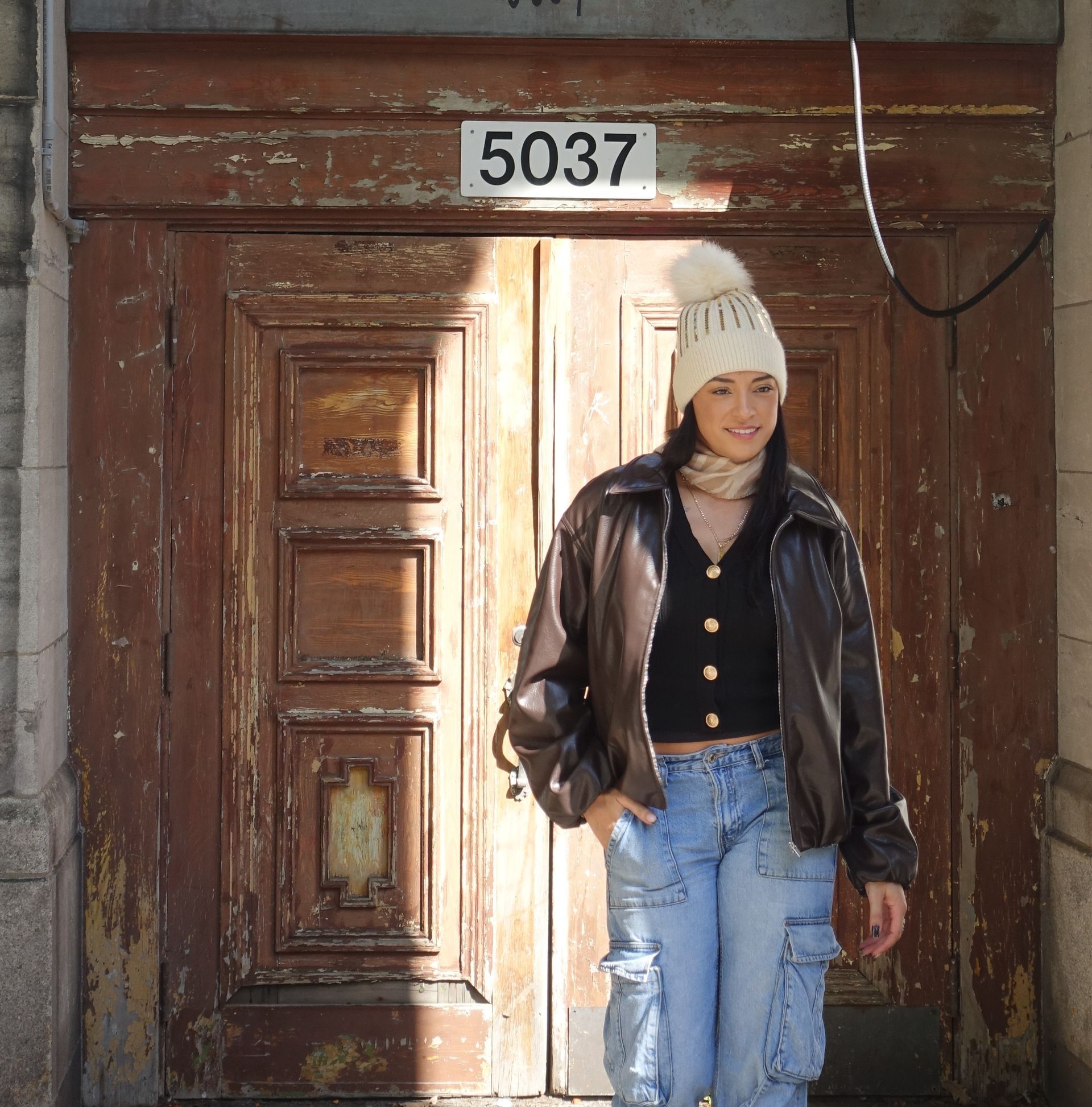Woman smiling in front of an old wooden door with the number 5037 above it. She wears a brown jacket and blue jeans.