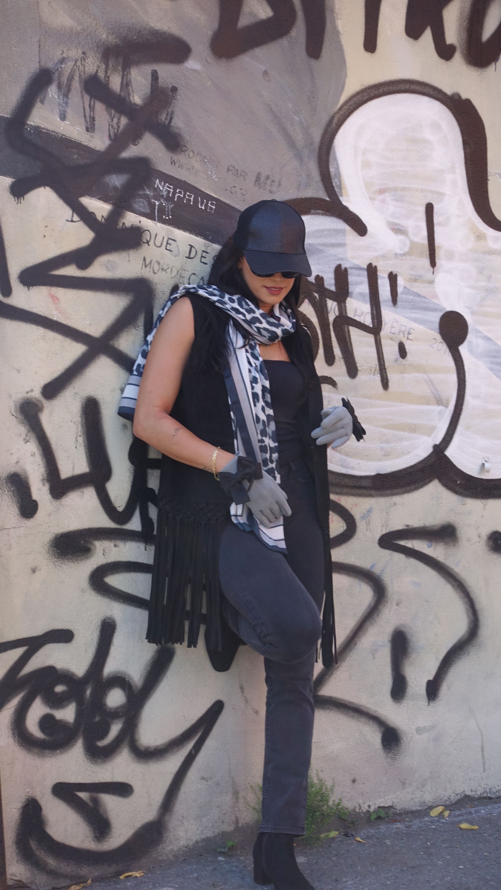 Person in black vest and cap, leaning against a graffiti-covered wall, wearing scarf and gloves.