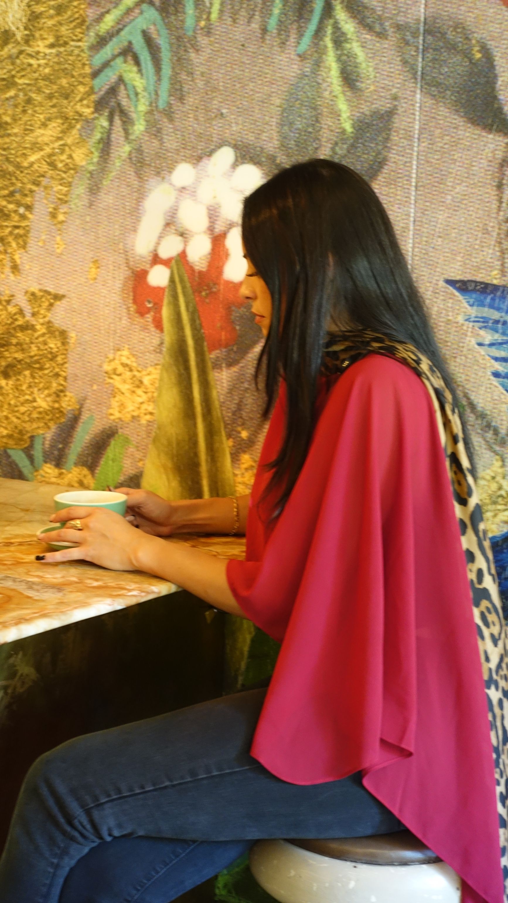 Woman in red top and jeans at a table, floral mural backdrop.