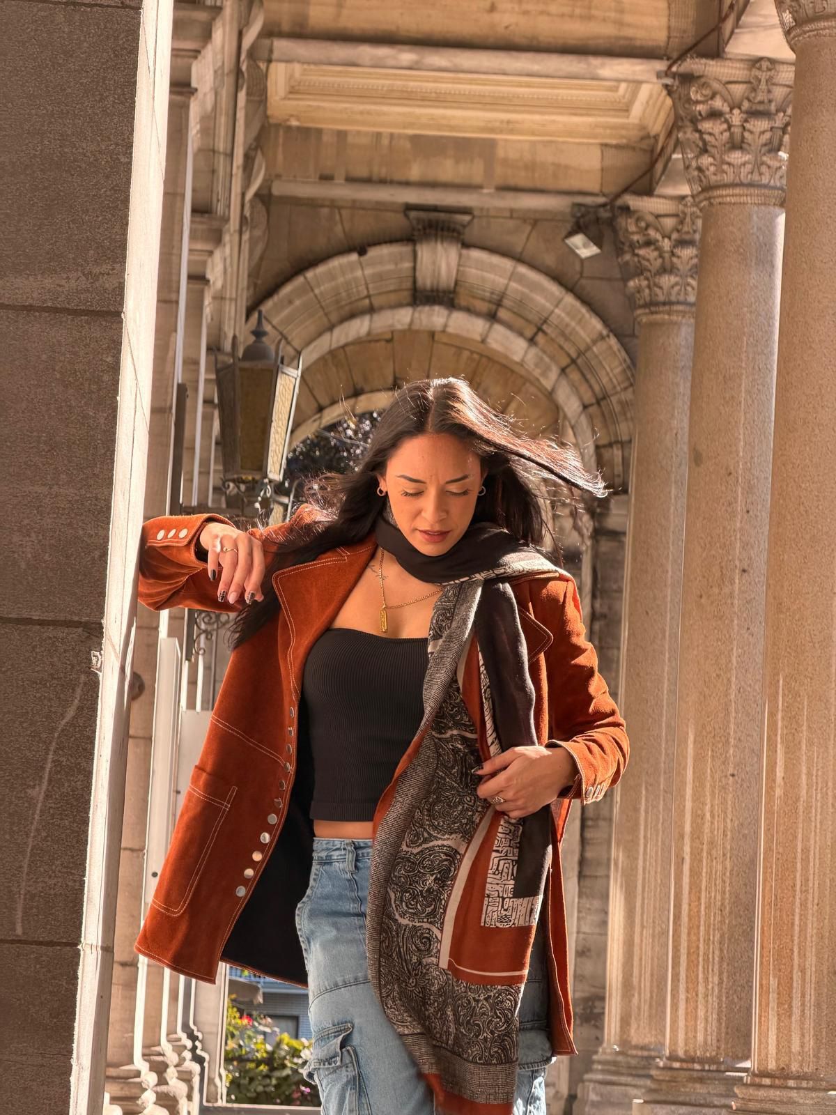 Woman in a brown coat and scarf adjusting her clothing under an arched walkway with pillars.