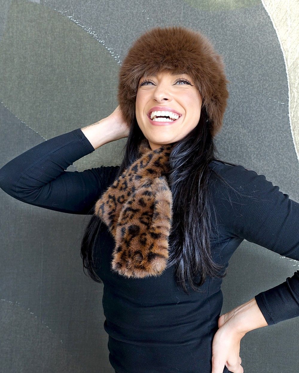 Woman smiling, wearing brown fur hat and leopard print scarf, black top, against a textured wall.