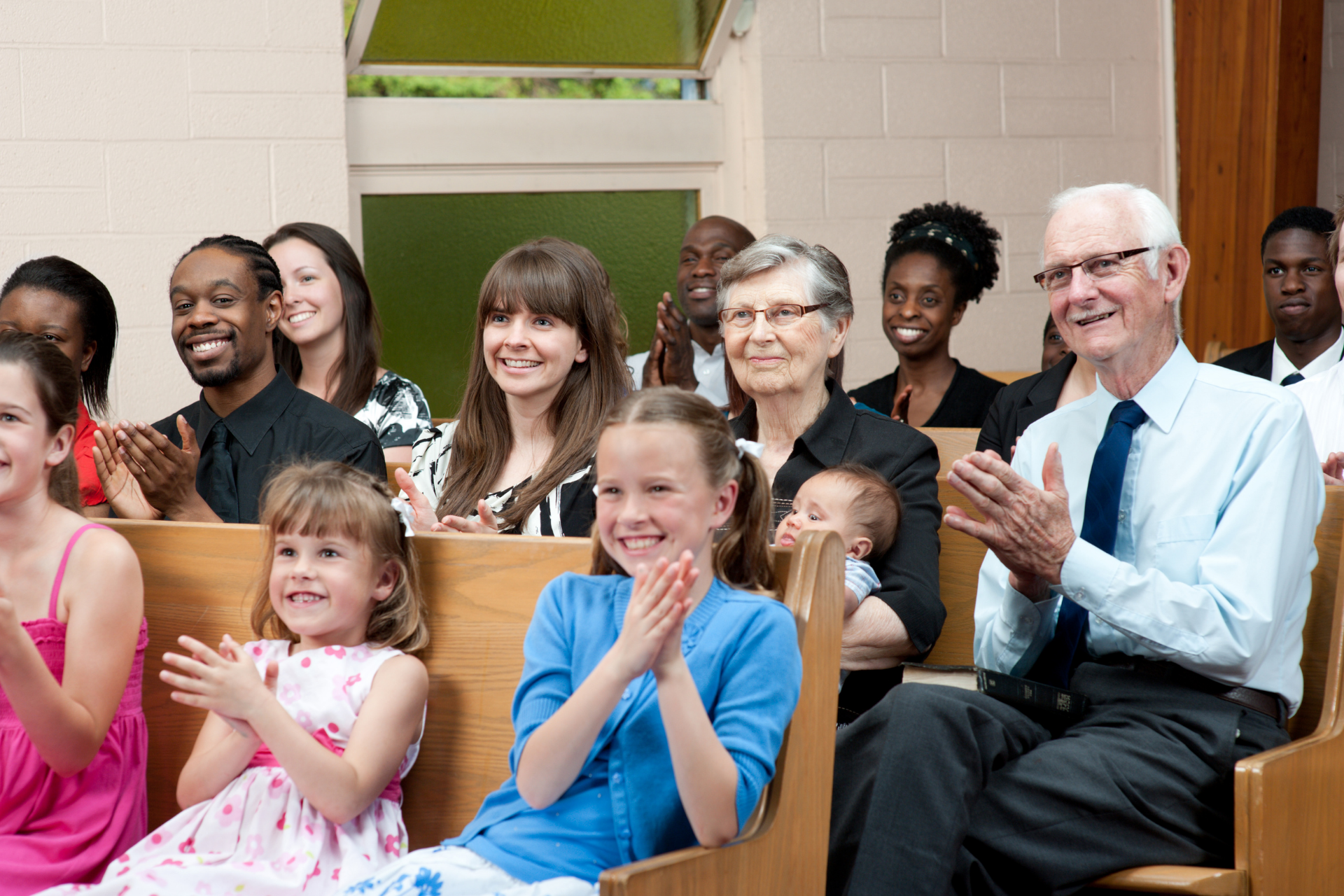 People clapping in a church. Pews filled with diverse individuals, smiling, some with children, inside a bright setting.