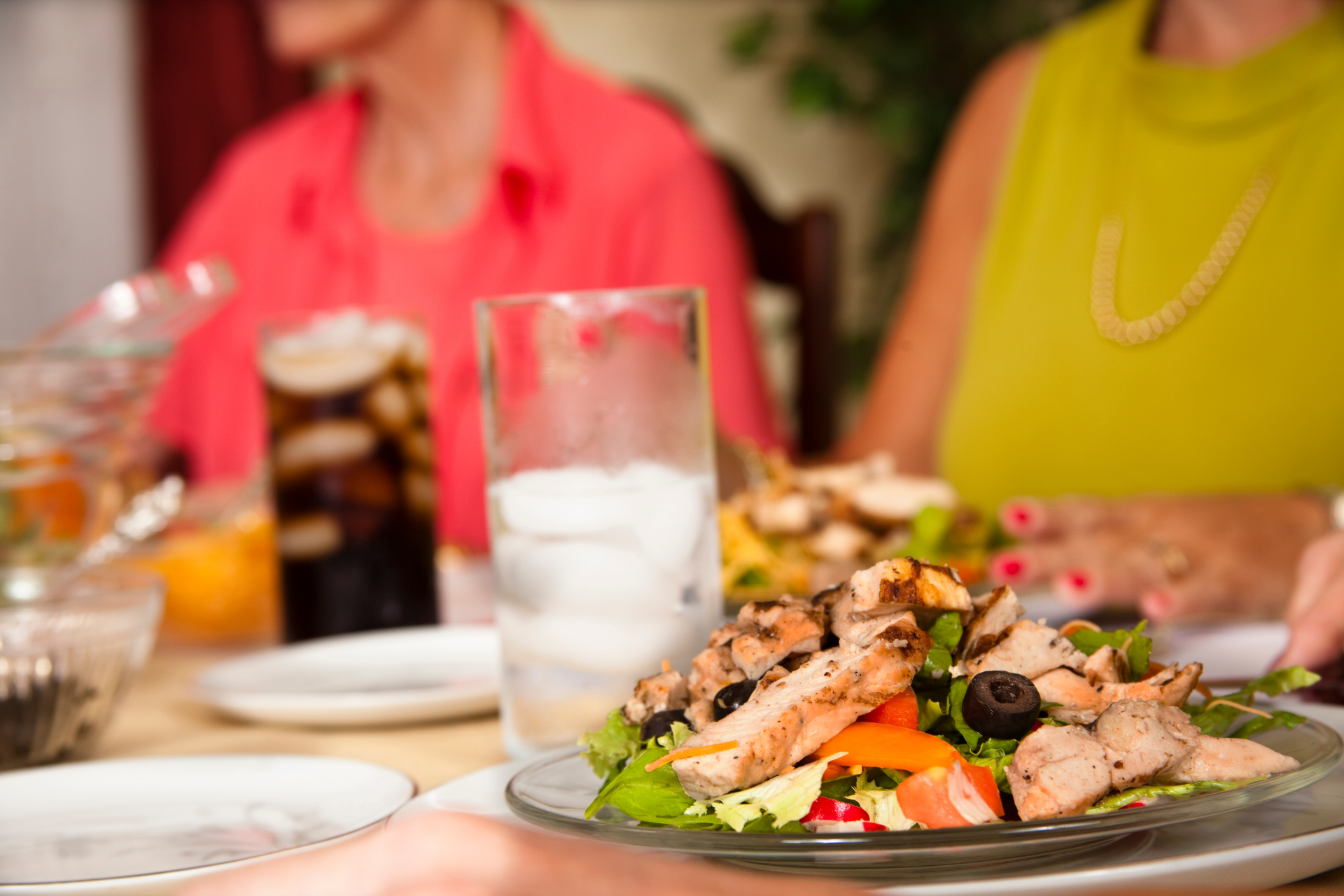 A salad with grilled chicken at a table with two people. One wears a yellow shirt, the other, a pink shirt.
