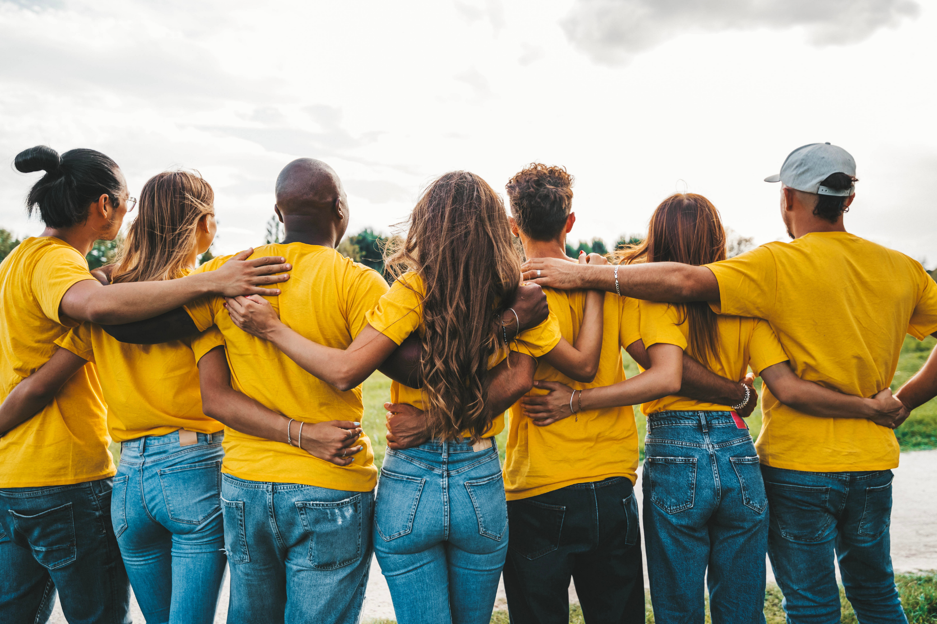 Group of people in yellow shirts and jeans with arms around each other, facing a sunny landscape.