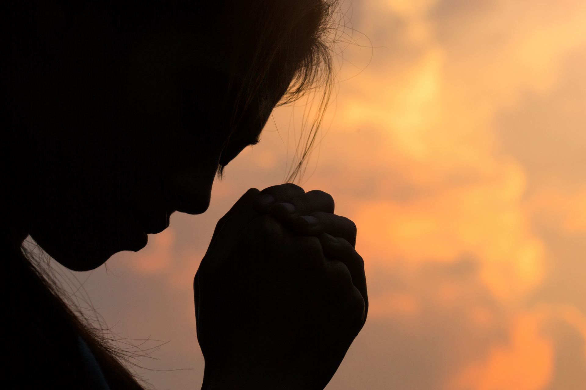 Silhouette of a person with hands clasped in prayer, set against a warm, orange-toned sky.
