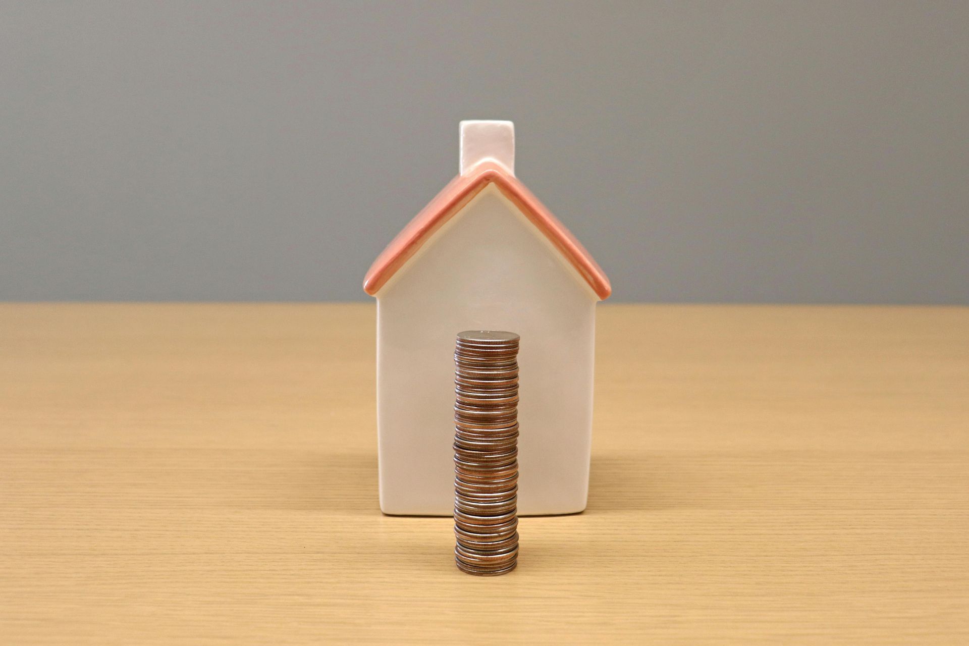Small white house model with orange roof and a stack of coins in front on a wooden table