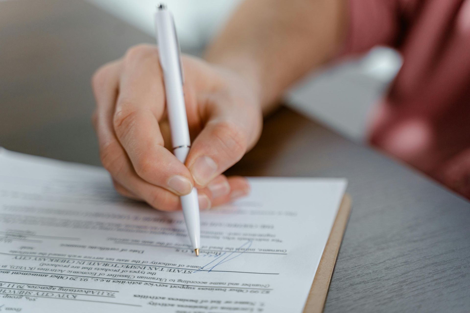 A hand holding a white pen, signing a document on a wooden table.