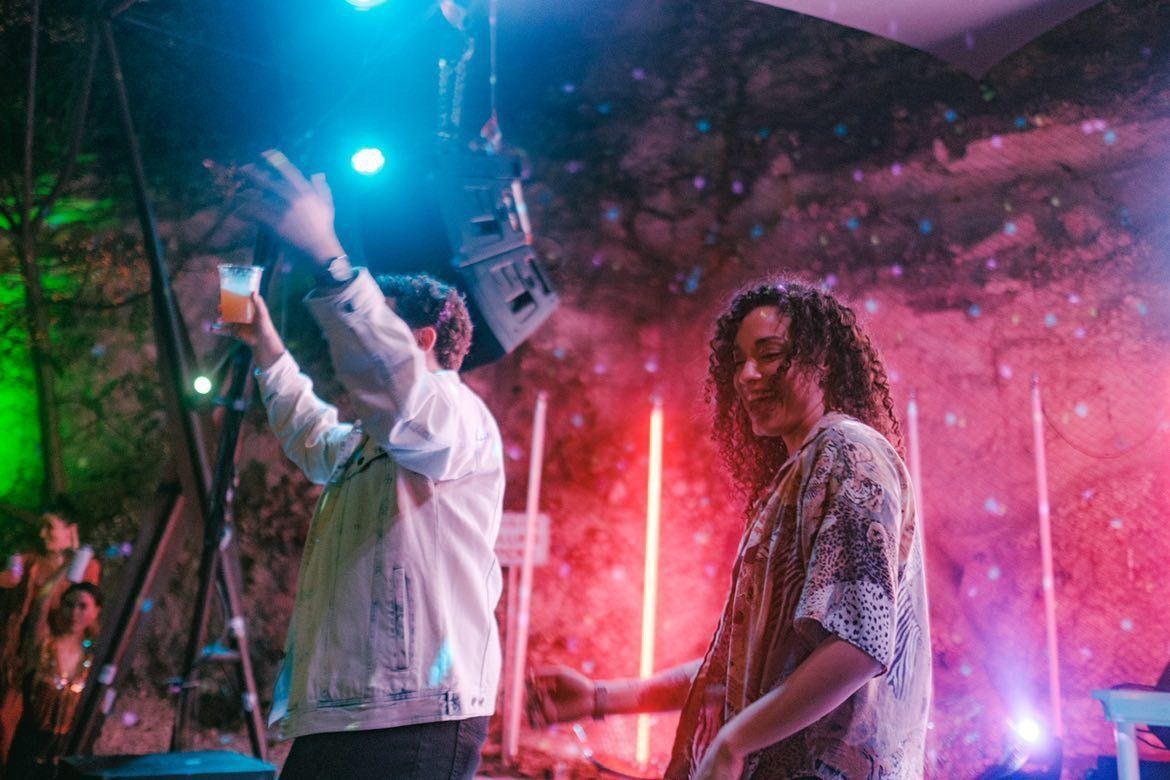 Concert attendees dance and drink to the music, backlit by neon event lights and stage equipment.