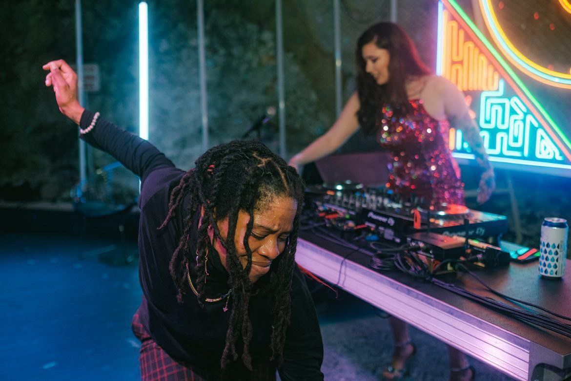 A woman dances in front of a DJ turn table while an artist spins music in a sequin dress.