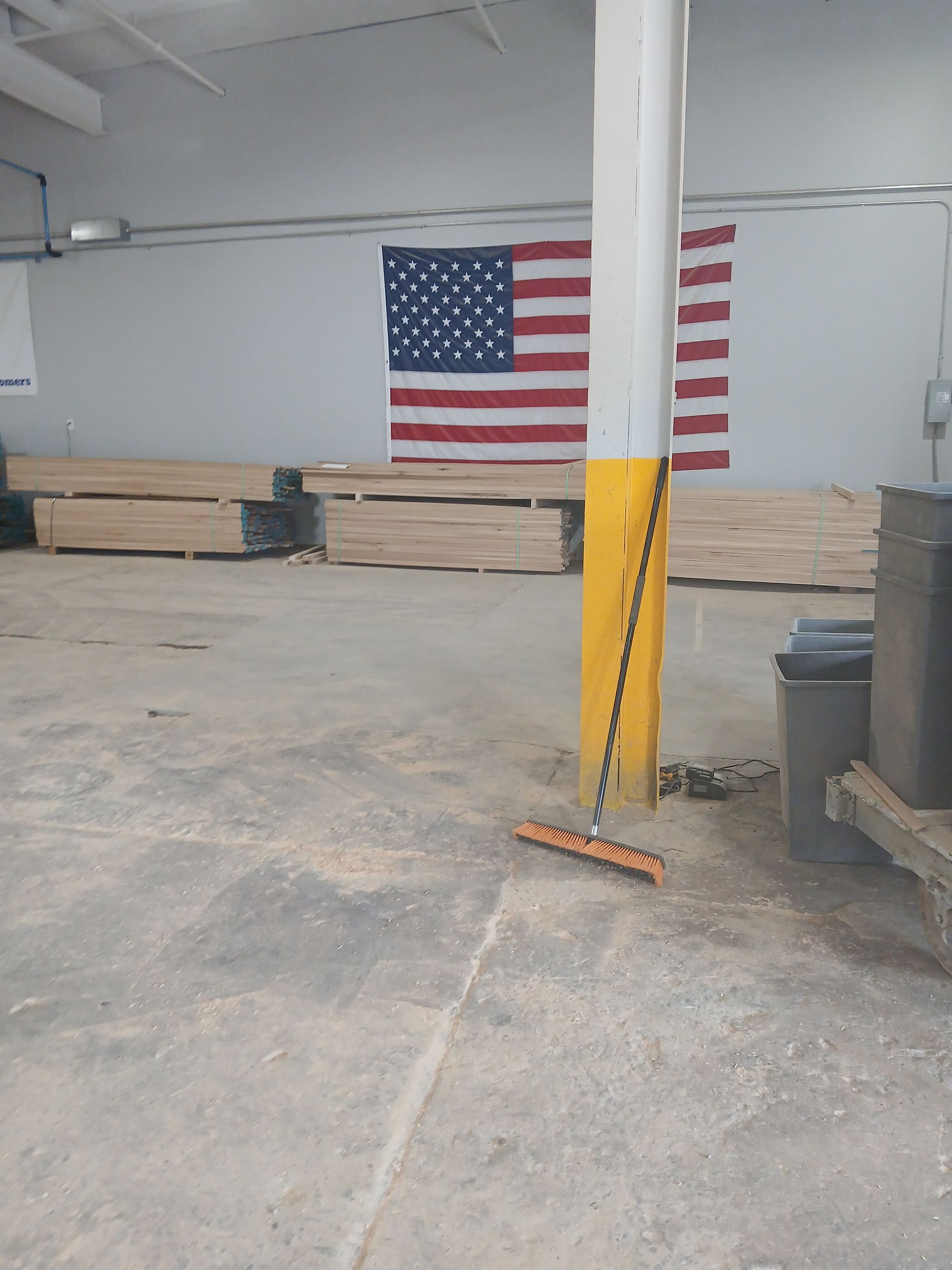 Warehouse interior with stacked lumber, a large American flag, and a broom leaning against a yellow pillar.
