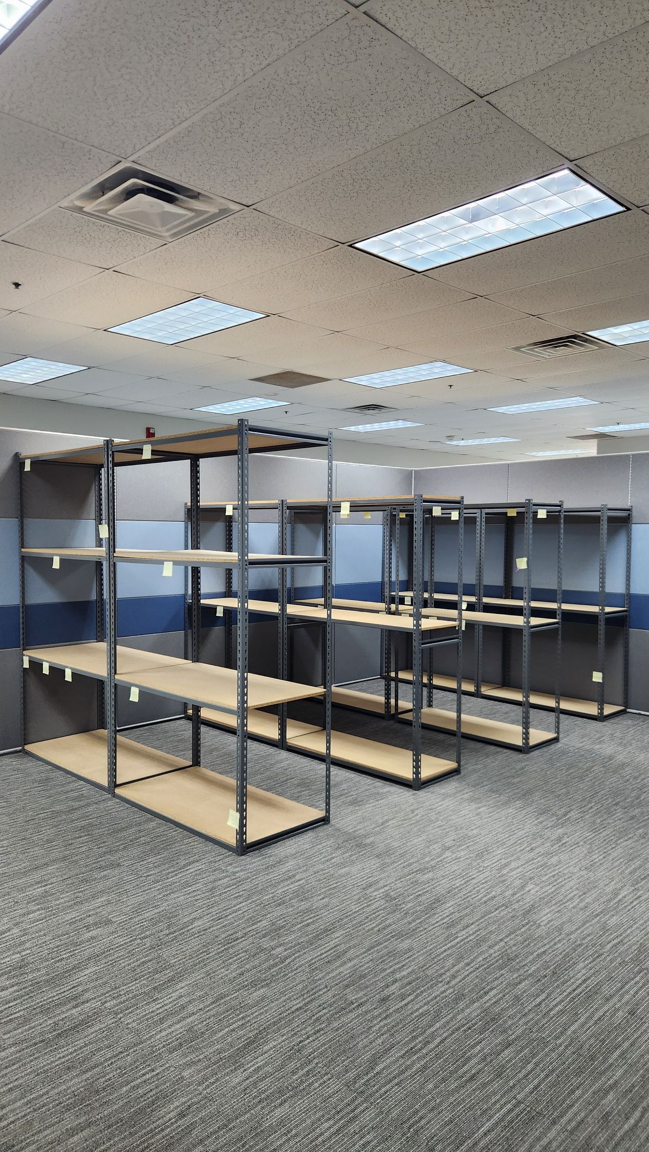 Empty office room with metal shelving racks and blue partitions under fluorescent ceiling lights.