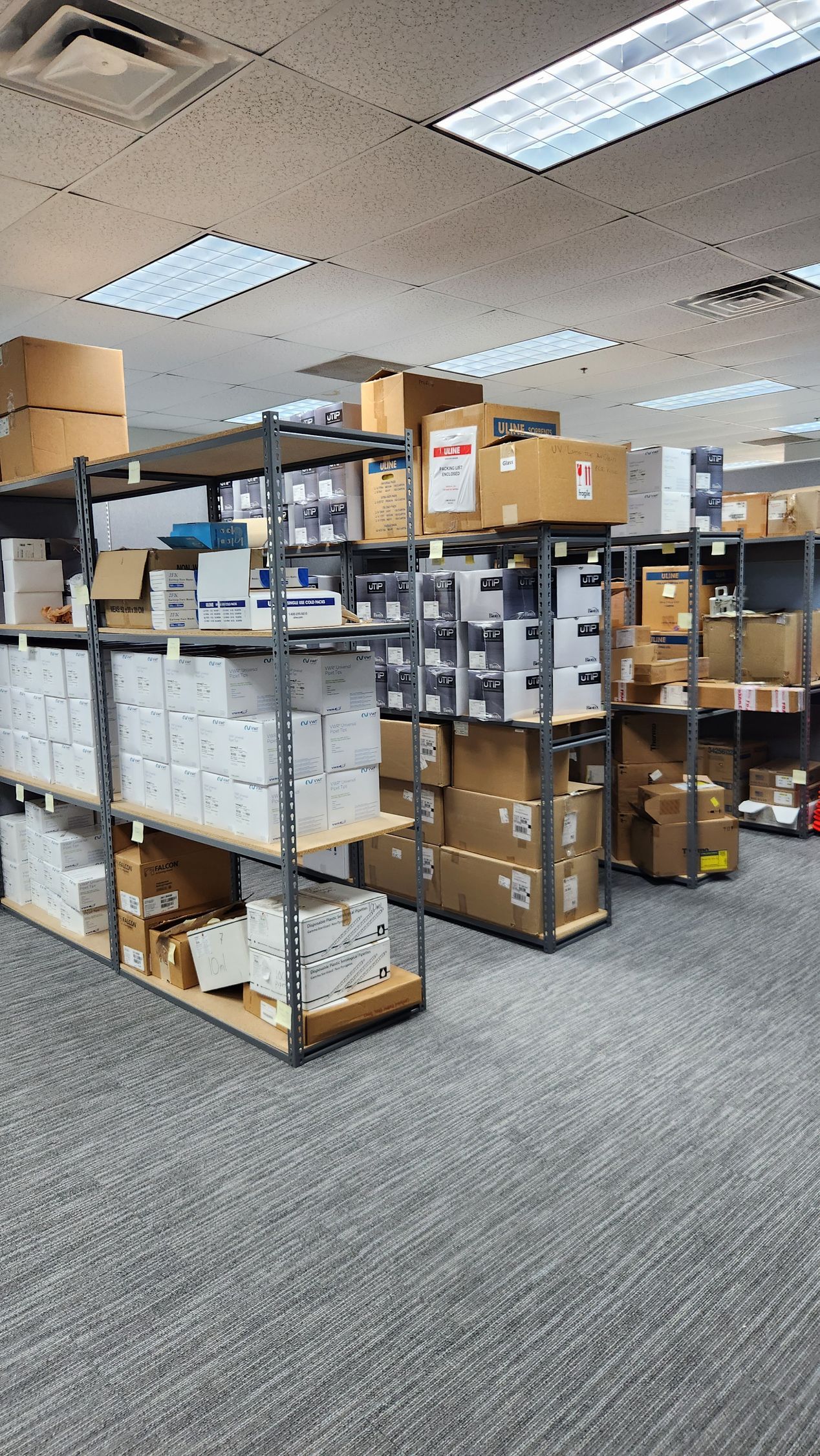 Stockroom aisle with shelves of labeled boxes and supplies under fluorescent ceiling lights.