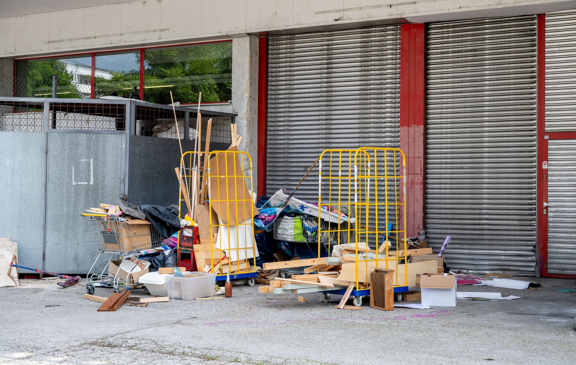 Construction materials and debris piled outside a closed storefront with metal shutters and red columns