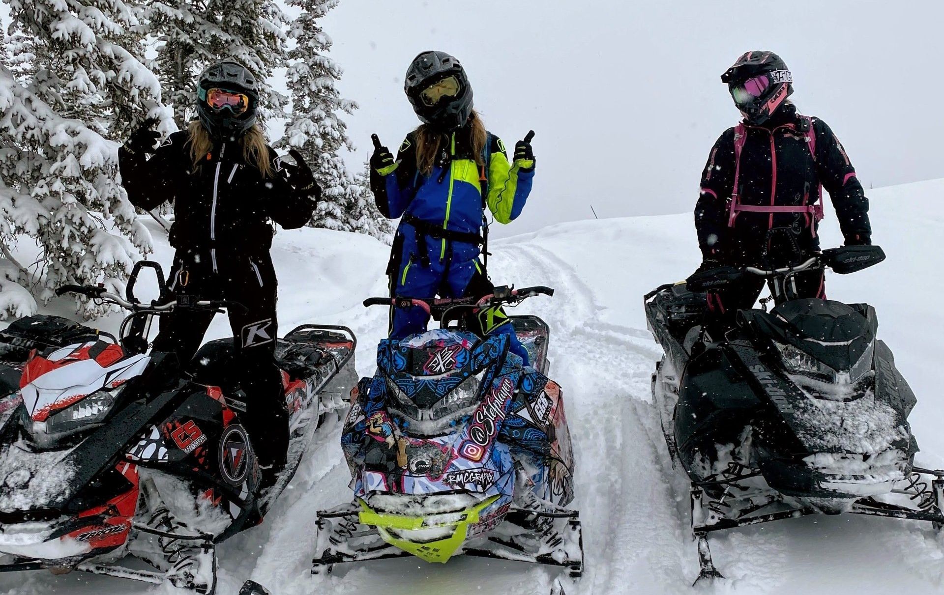 three female snowmobile riders posing in the snow on their snowmobiles