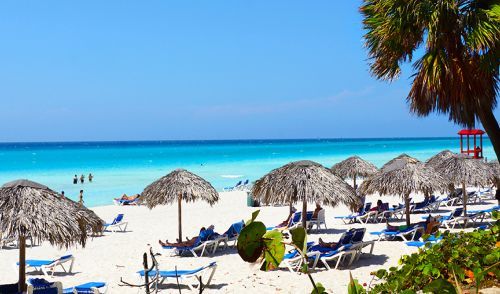 Beach scene with white sand, turquoise water, palm trees, thatched umbrellas, and people relaxing.