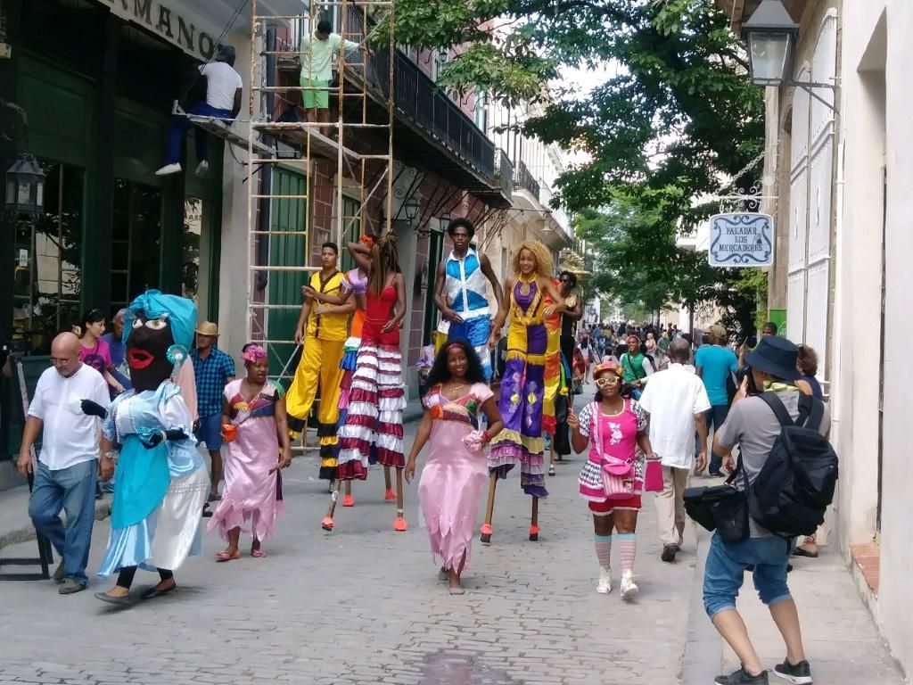 Street performers on stilts parade down a Havana street, entertaining onlookers; buildings and scaffolding in the background.