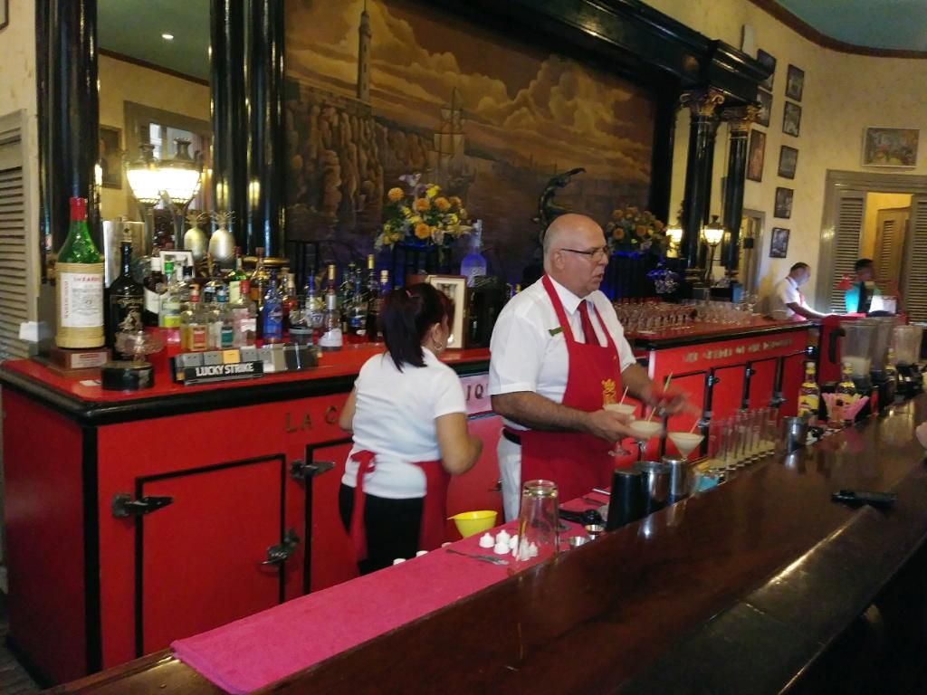 Bartenders preparing drinks behind a red bar. One wears a red apron. The setting is indoors, with a mural backdrop.