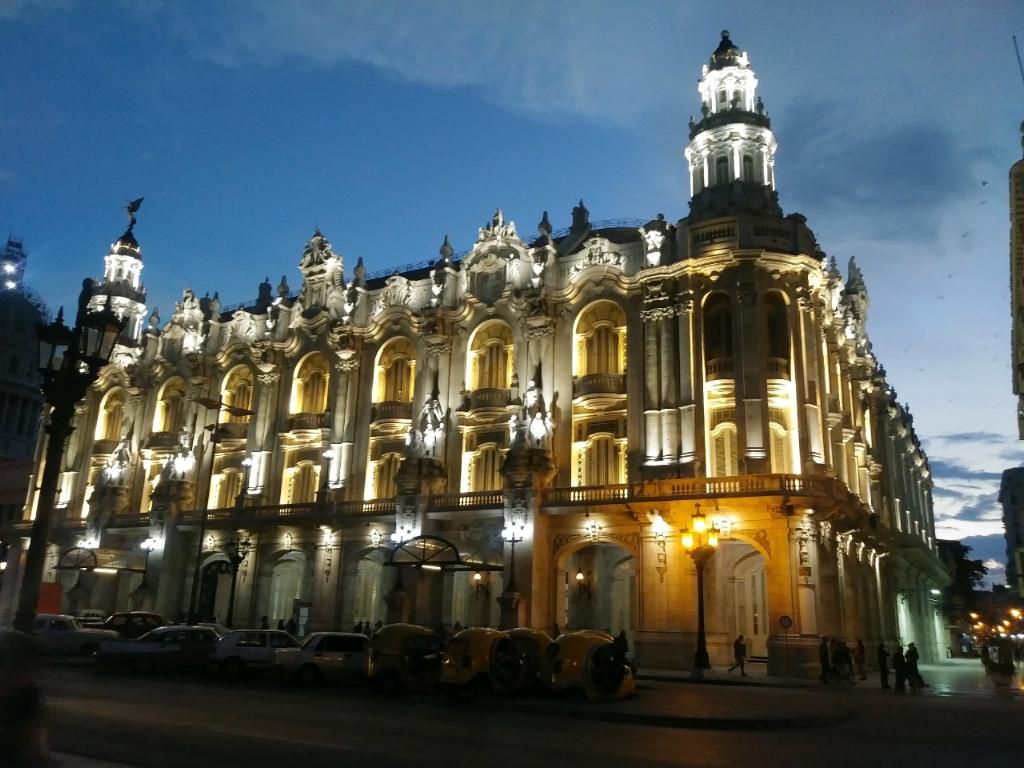 Illuminated ornate building at dusk. Facade with many windows, tower, and streetlights.