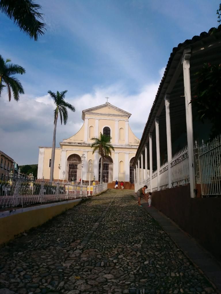 Cobblestone street leading to a light-colored church with palm trees and a colonnaded building.