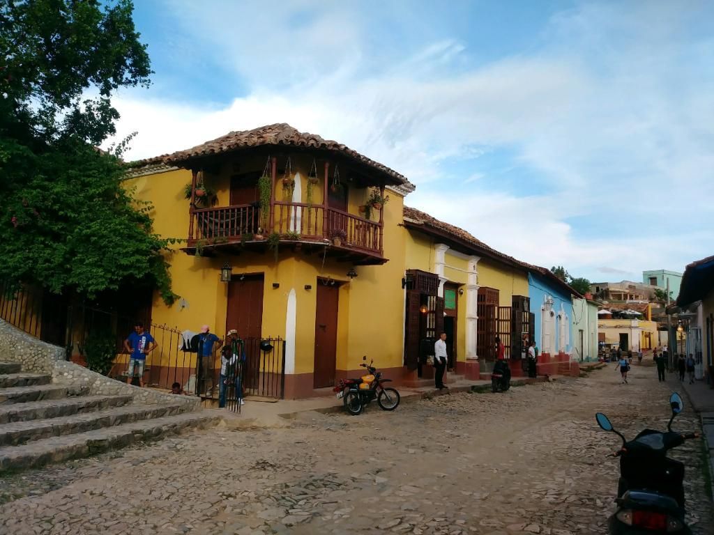 Cobblestone street in Trinidad, Cuba with yellow and blue buildings, people, and motorcycles.