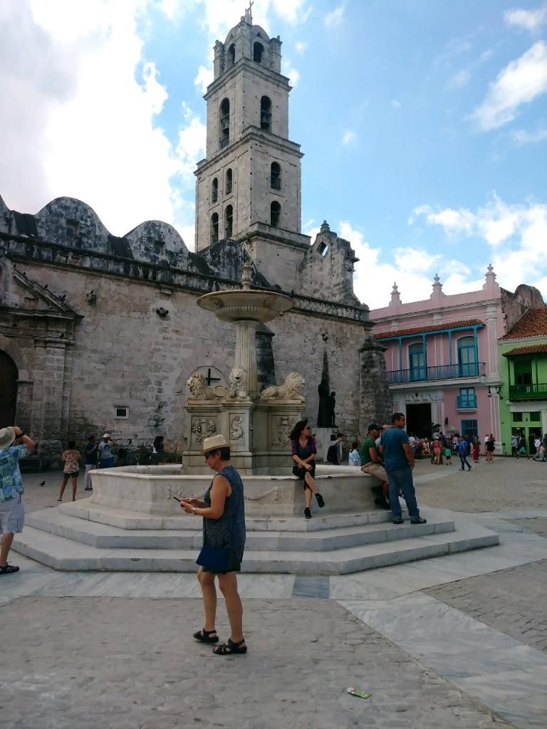 A plaza in Havana with a fountain, church tower, and colorful buildings; people are walking and sitting.