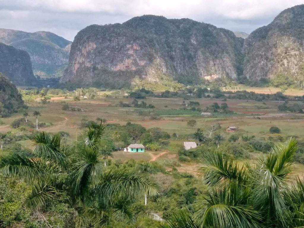 Valley landscape with lush green vegetation, small buildings, and large, rocky hills under a cloudy sky.