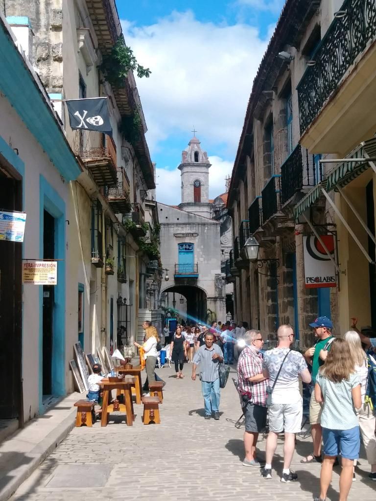 Narrow street in Havana, Cuba, lined with colorful buildings. People walking toward archway under a tower.