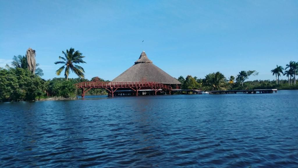 A large thatched-roof building on stilts over water, connected by a wooden walkway. Blue sky.