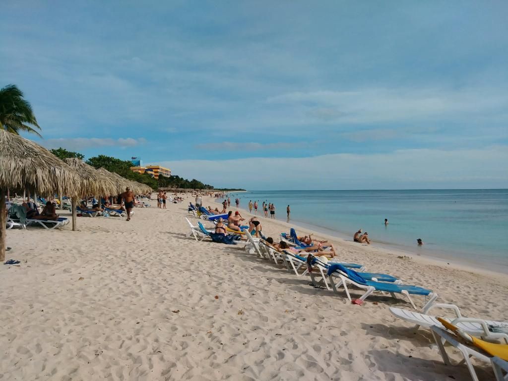 Beach with white sand, blue water, and lounge chairs under thatched umbrellas; people are relaxing.