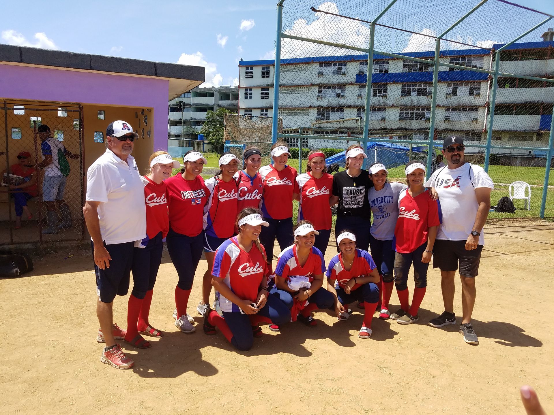 Softball team in red and blue uniforms poses on a field with two men. Buildings in the background.
